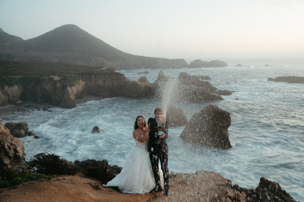 A bride and groom stand on a rocky cliff above the ocean, laughing as champagne sprays into the air.
Waves crash below them, and the open sea stretches behind, creating a joyful, wild moment.