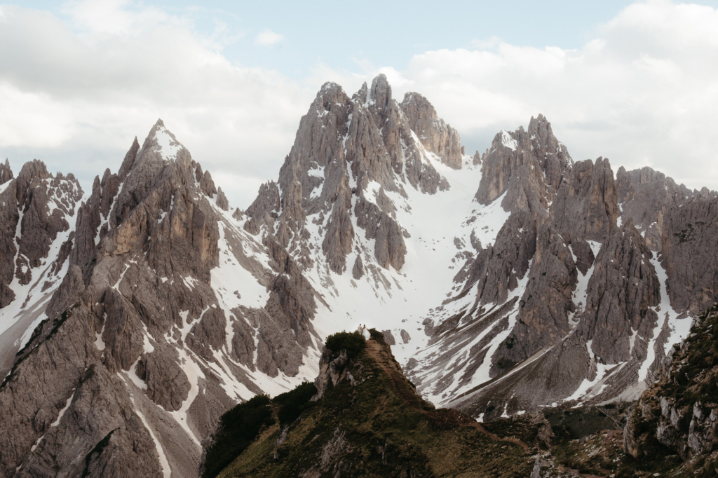 A vast mountain landscape with jagged, rocky peaks dusted with patches of snow.
In the distance, a tiny couple stands on a narrow ridge, surrounded by dramatic cliffs and open sky.