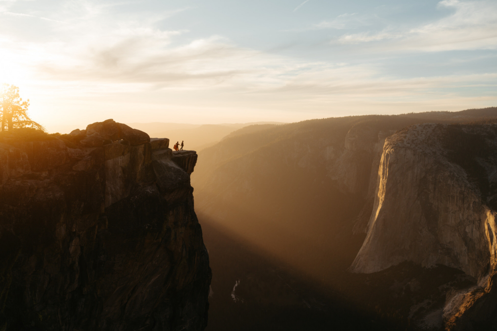 Two small figures stand on the edge of a high cliff overlooking a deep valley at sunset.
Golden light fills the scene, with layered mountains fading into the distance.