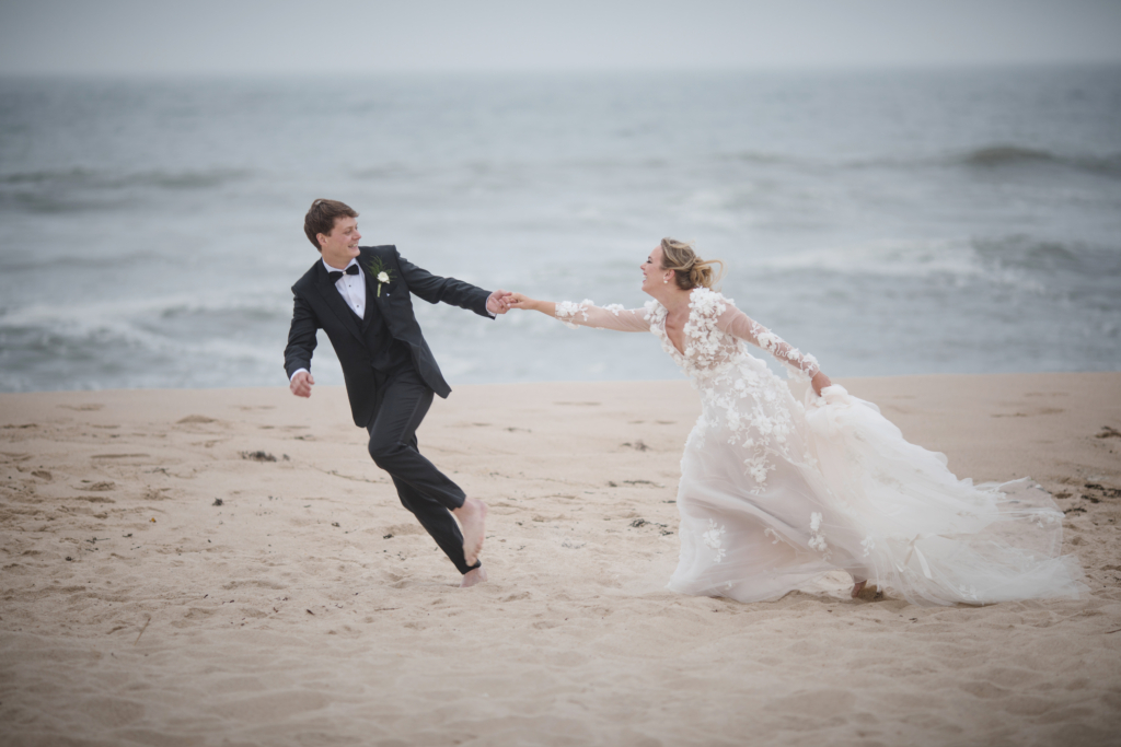 A bride and groom run hand in hand along a sandy beach, smiling as the wind lifts the bride’s dress. The ocean behind them adds a sense of movement and carefree joy.