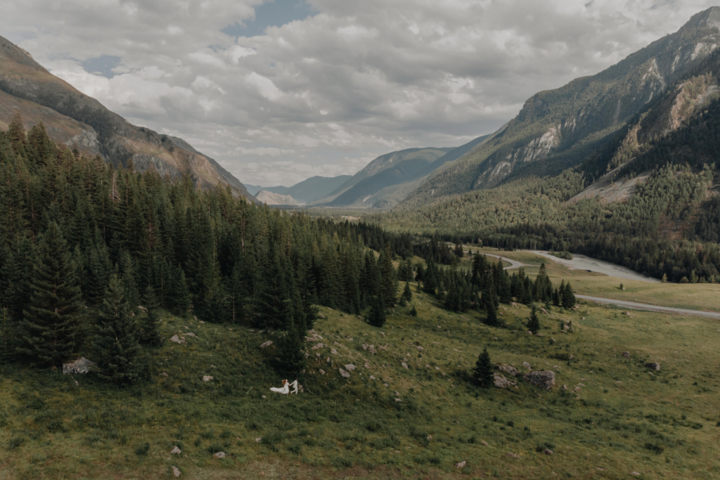 A wide mountain valley with dense pine forests, green meadows, and a winding river. Two small figures in white stand together in the grass, surrounded by towering mountains under a cloudy sky.