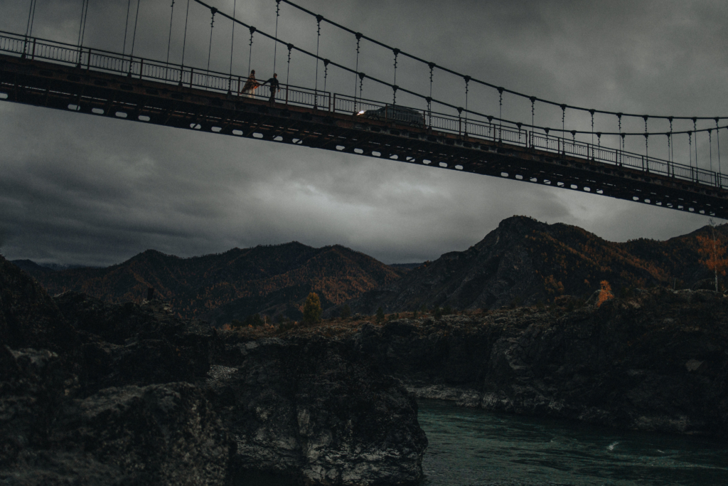 A suspension bridge stretches across the top of the image, with two small figures standing on it. Below, dark rocky terrain and a river cut through a mountainous landscape. Heavy clouds fill the sky, creating a dramatic, moody atmosphere.