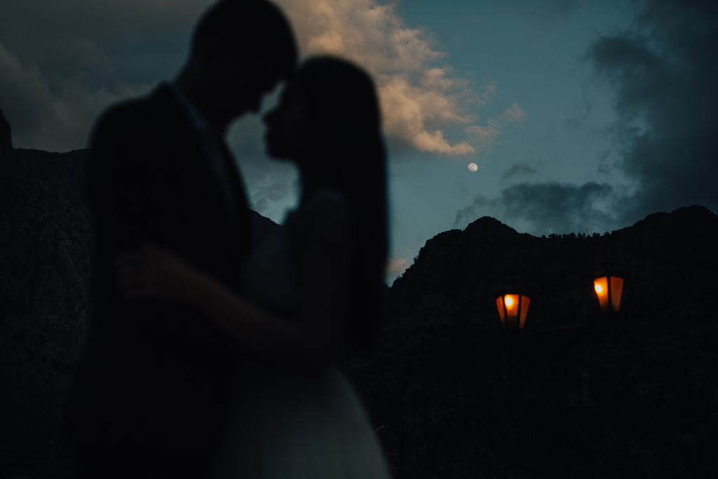 A silhouetted couple stands close together at night, their faces nearly touching. Behind them, dark mountains rise under a cloudy sky with a bright moon visible. Two warm streetlights glow in the distance, adding a quiet, intimate contrast to the night scene.