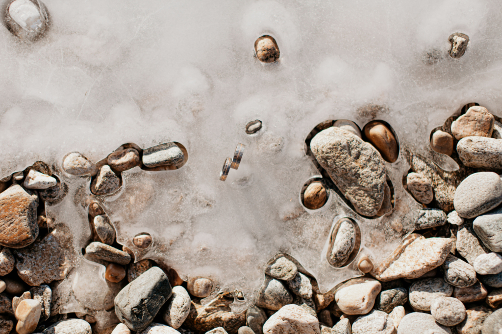 A close-up photograph shows small stones and pebbles trapped beneath a thin layer of ice.
The ice appears cloudy and cracked in places, creating soft white patches that contrast with the warm browns and grays of the rocks below.
The textures of smooth pebbles, rough stones, and frozen water blend together, giving a quiet, fragile feeling, as if nature is paused in mid-winter.