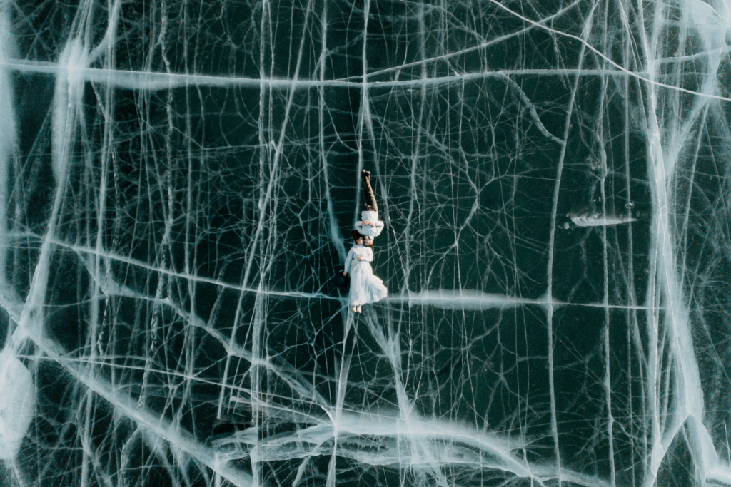 A couple lies side by side on a frozen lake, seen from directly above. The dark ice beneath them is crossed with white cracks that form a web-like pattern, while their light clothing contrasts sharply against the textured surface, creating a quiet, surreal sense of stillness.