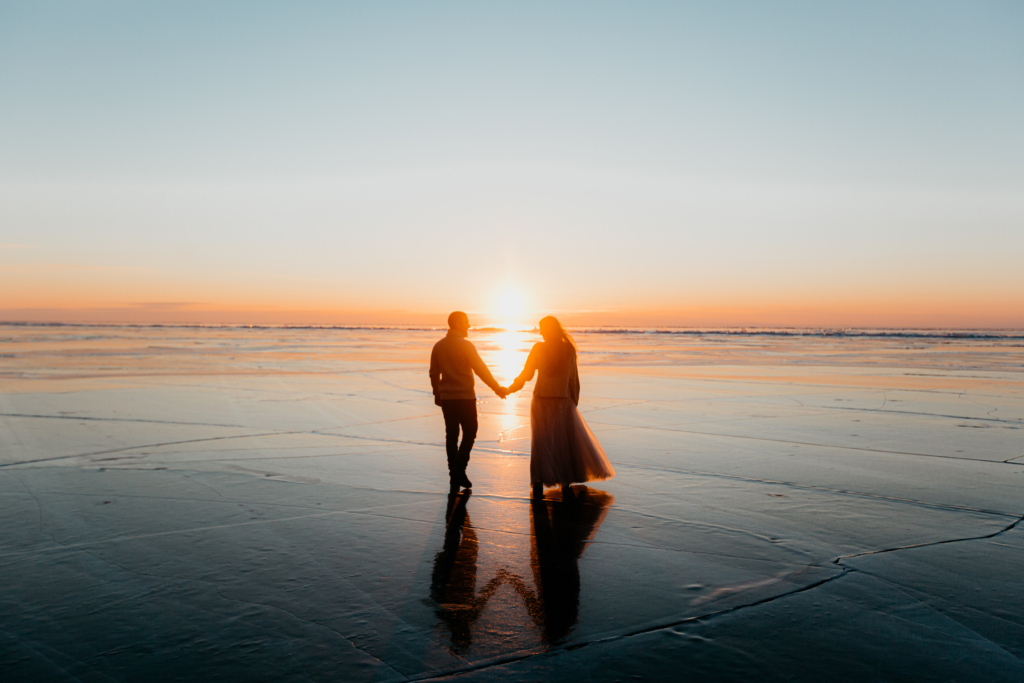 A couple walks hand in hand across wet sand at the edge of the ocean during sunset. The low sun glows between them, casting long reflections on the water. The sky fades from warm orange near the horizon to soft blue above, creating a peaceful, intimate scene.