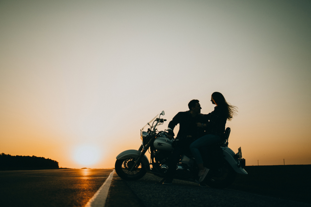 A couple sits on a motorcycle stopped on an open road at sunset. They face each other closely, silhouetted against a glowing orange sky. The long road stretches into the distance, creating a quiet, cinematic sense of freedom and intimacy.
