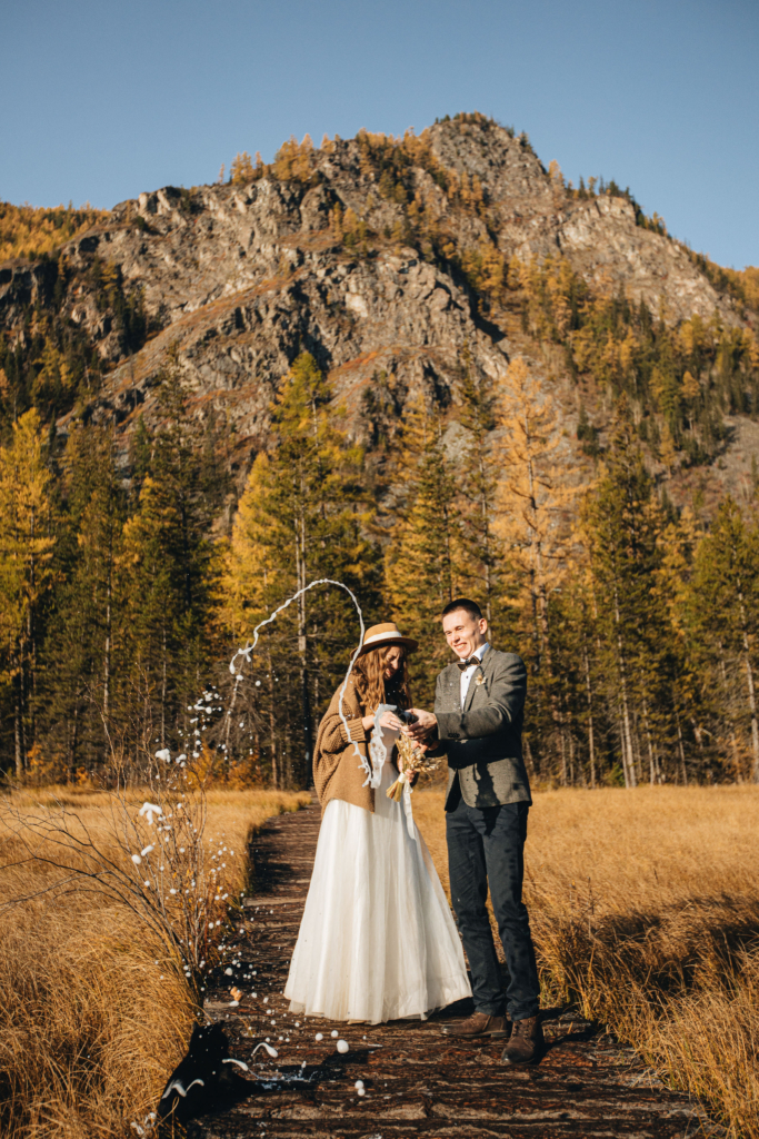 A couple stands on a narrow wooden path in a golden autumn landscape. They pop a bottle together, sending liquid splashing into the air. Tall trees and a rocky mountain rise behind them, creating a joyful, celebratory moment in nature.