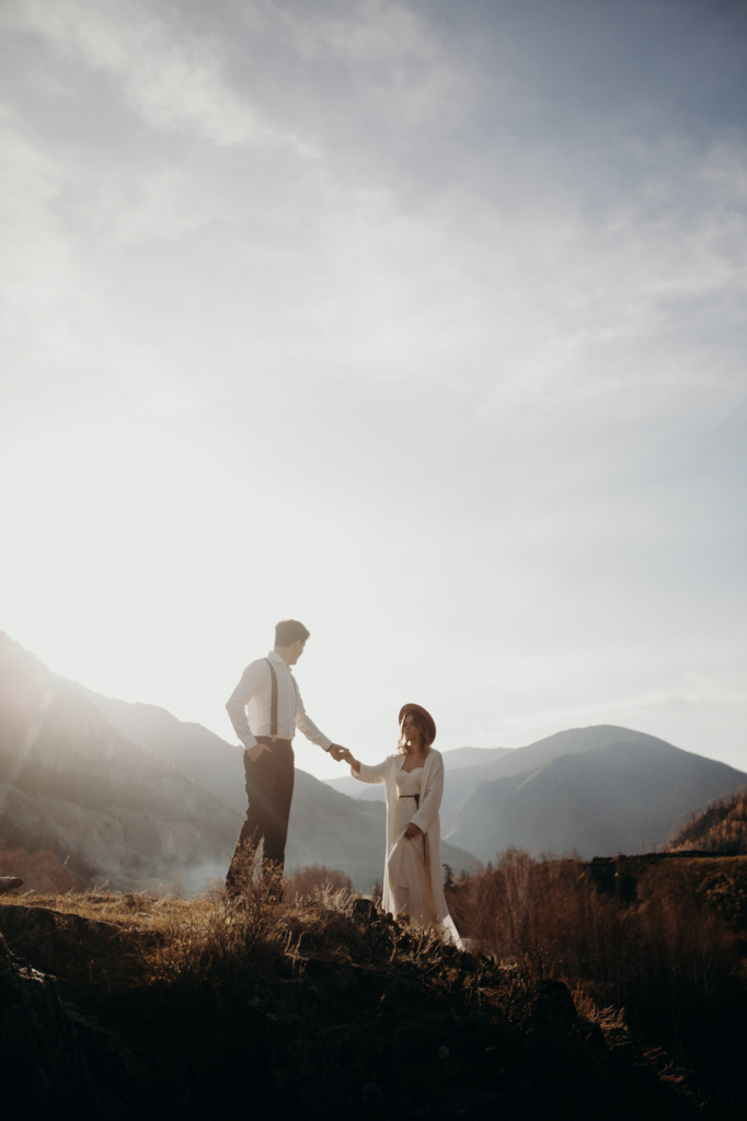 A couple stands on a rocky hillside, holding hands against a bright sky. The man wears suspenders and a white shirt, the woman a long light dress and a hat. Mountains fade into the background, and soft sunlight creates a calm, intimate atmosphere.