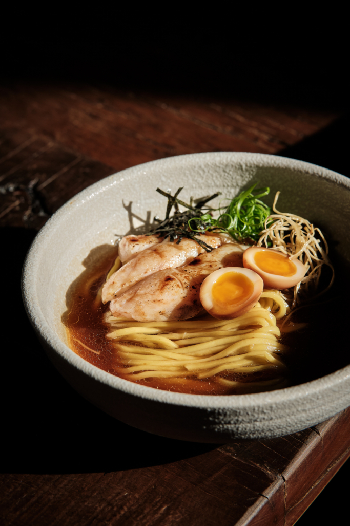 A bowl of noodles with meat and egg, photographed on a dark background.