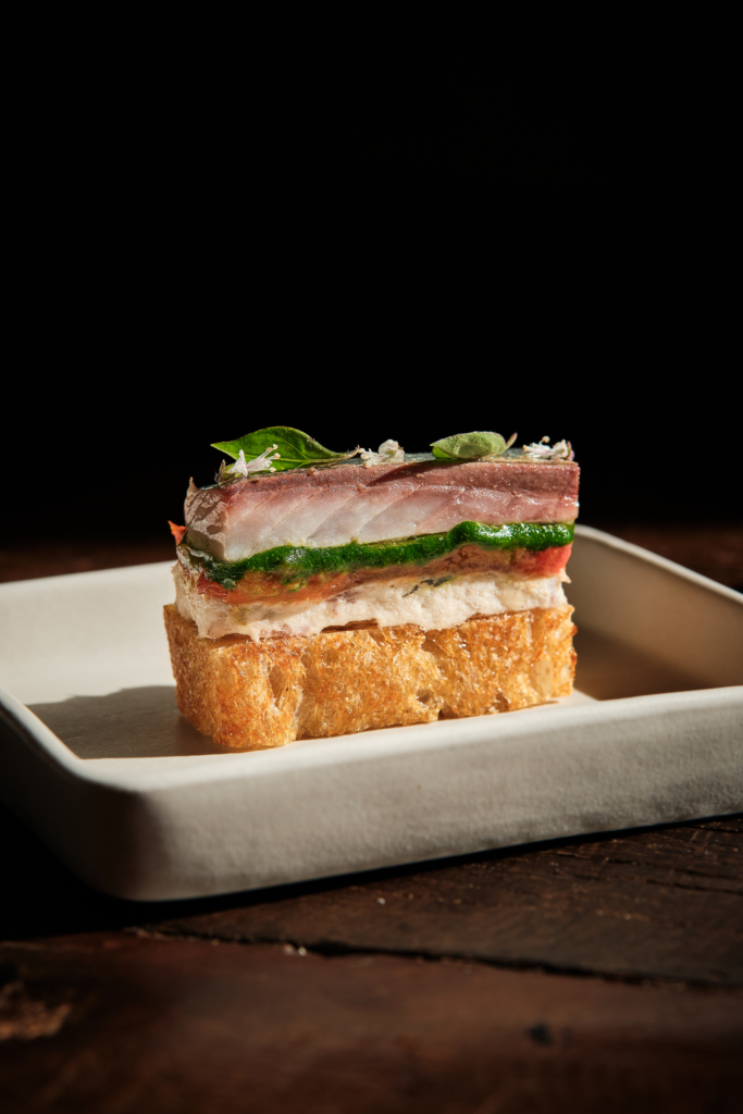A layered piece of food served on bread, photographed on a dark background.