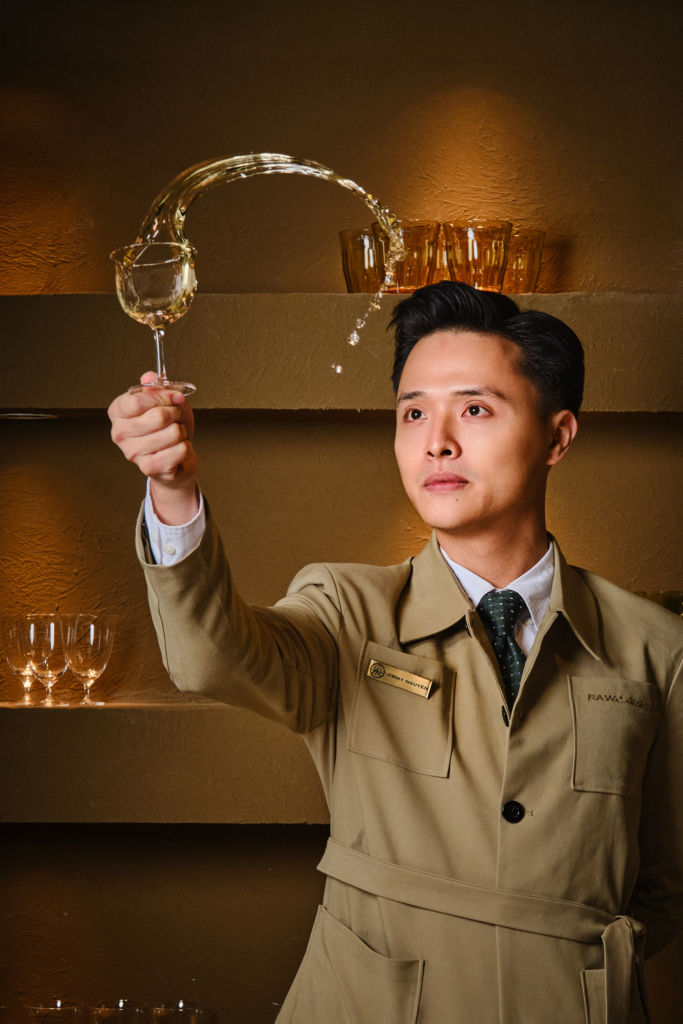 A well-dressed man stands in front of warm, golden shelves filled with glassware. He is holding a wine glass at arm’s length while pouring a clear liquid into it in a high arc, creating a curved stream of liquid in the air. He wears a beige jacket with a name tag and a dark tie, and looks focused and calm. The background is softly lit, with multiple empty glasses arranged on the shelves behind him.