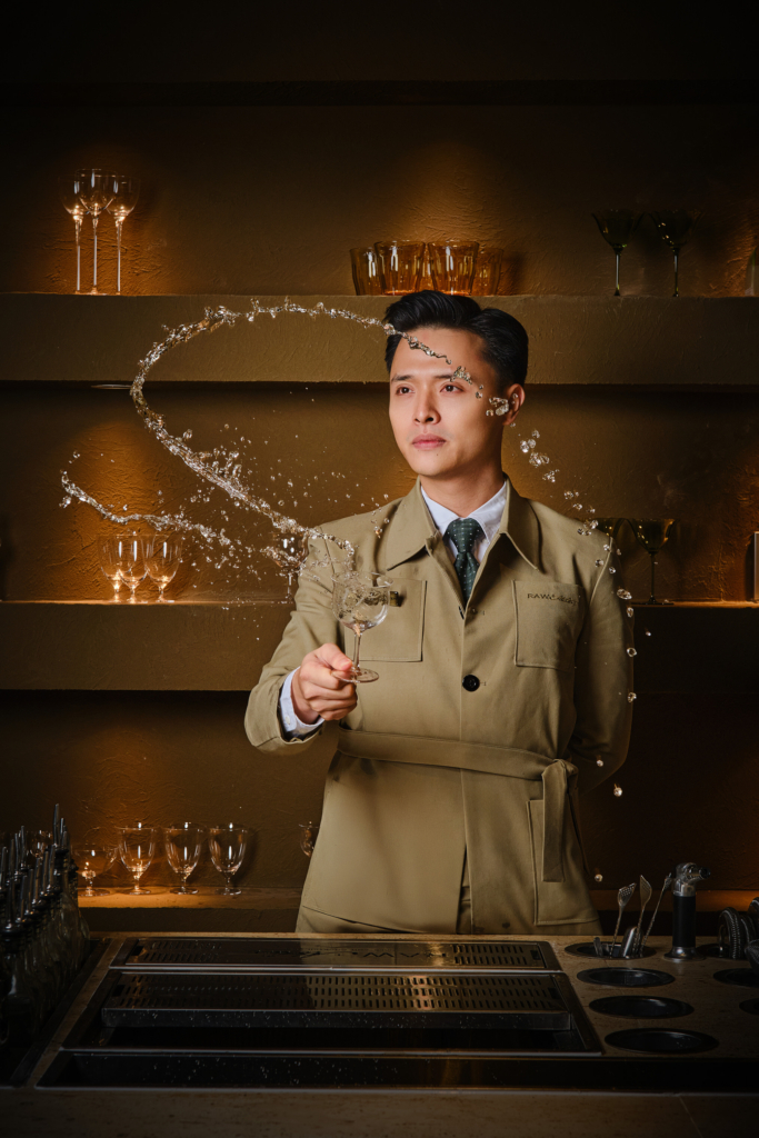 A well-dressed man stands in front of warm, golden shelves filled with glassware. He is holding a wine glass at arm’s length while pouring a clear liquid into it in a high arc, creating a curved stream of liquid in the air. He wears a beige jacket with a name tag and a dark tie, and looks focused and calm. The background is softly lit, with multiple empty glasses arranged on the shelves behind him.