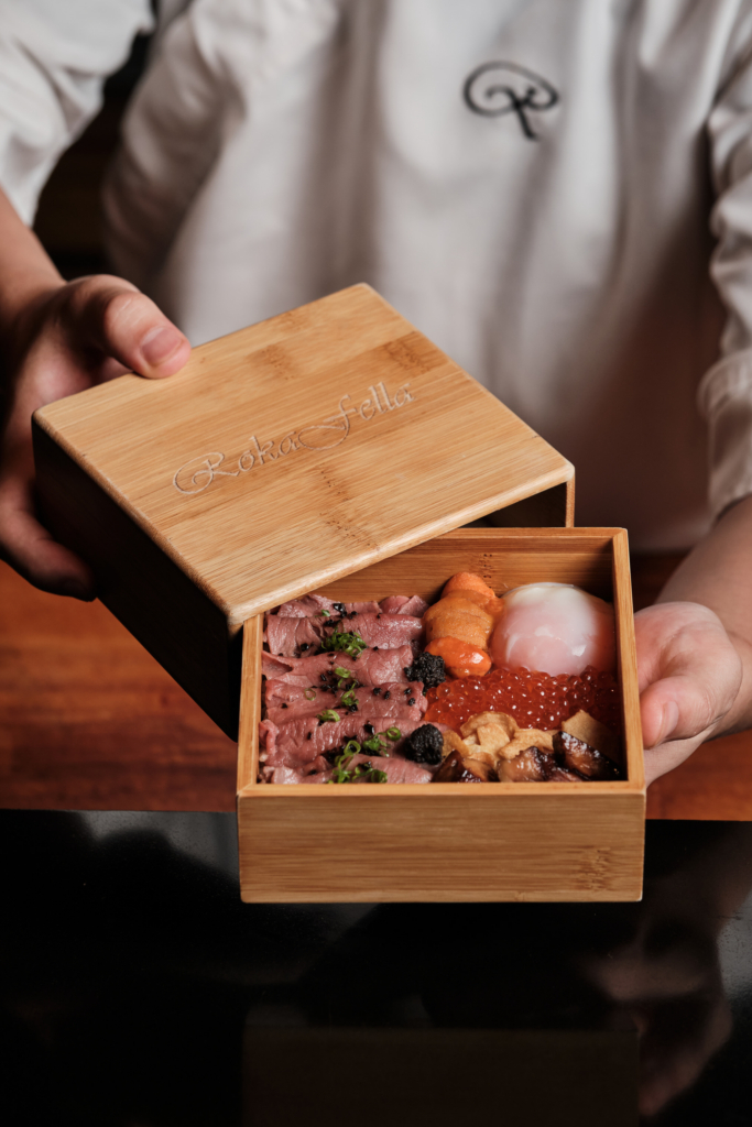 A wooden box with assorted food items, held in two hands, photographed on a dark surface.
