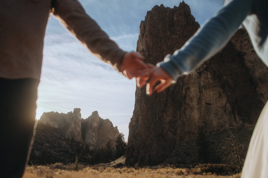 Two people hold hands in the foreground, seen from a low angle. Their figures are partially out of frame, with tall rocky cliffs rising behind them. Warm sunlight fills the scene, creating a sense of connection and scale within a vast natural landscape.