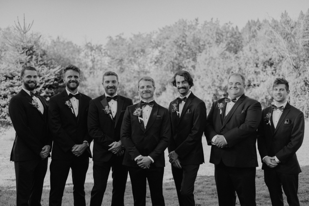 A black-and-white photograph shows eight men standing side by side outdoors, dressed formally in dark suits with bow ties and boutonnières.
They are arranged in a relaxed line, hands loosely clasped, smiling softly toward the camera.
Behind them is a bright, slightly blurred background of trees and foliage, suggesting a calm outdoor setting, possibly before a formal celebration or ceremony.