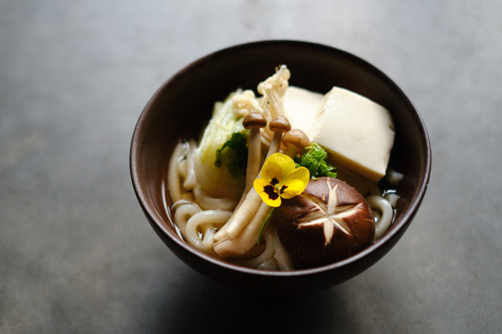 A close, slightly top-down view of a dark bowl filled with thick white noodles in clear broth. On top are several types of mushrooms, including a large brown mushroom with a star-shaped cut on its cap and a cluster of small beige mushrooms. A bright yellow edible flower sits in the center. There are also pieces of pale tofu, chopped green herbs, and light-colored vegetables arranged neatly. The bowl is placed on a gray surface with soft natural light.