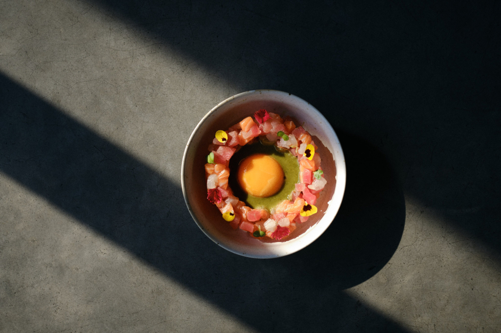 A top-down view of a small white bowl placed on a dark surface. Inside the bowl is a circular arrangement of finely chopped raw fish in shades of pink and orange, forming a ring. In the center of the ring is a bright orange egg yolk sitting on a pool of green sauce. Small edible flowers and herbs are scattered on top. Strong diagonal shadows fall across the surface, creating a dramatic contrast between light and dark.