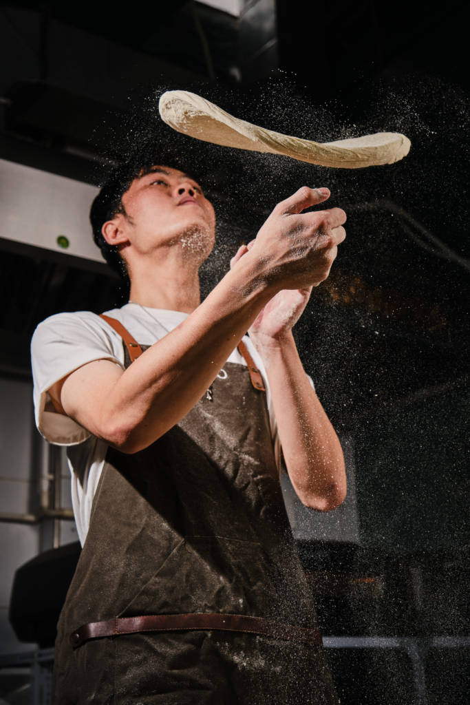 A chef tossing dough in the air with flour particles visible.