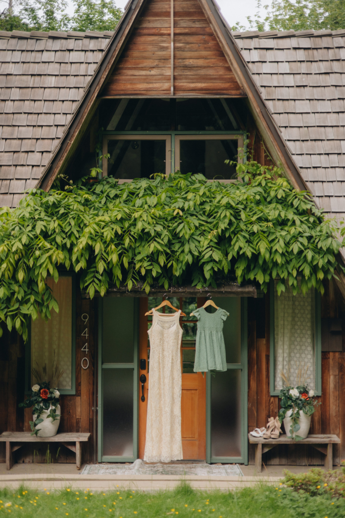 A small wooden cottage is covered with green vines around the entrance. Two dresses hang on the front door: a long white lace dress and a shorter green dress. Flower pots and shoes are placed on benches beside the doorway, creating a calm, intimate wedding-day atmosphere.