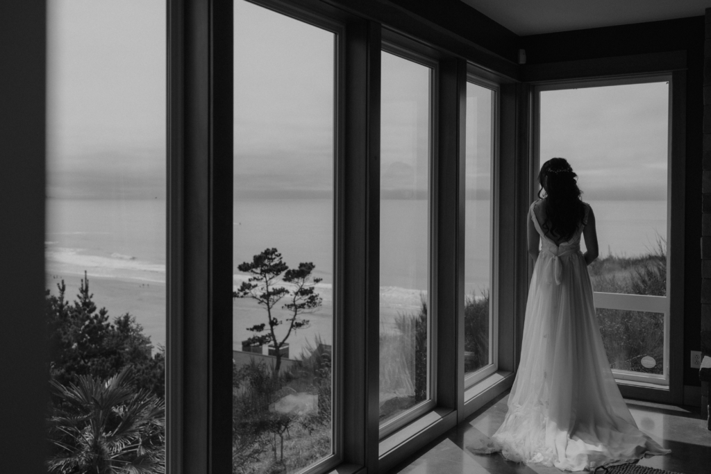 A woman in a long white dress stands with her back to the camera inside a room with large floor-to-ceiling windows. Outside, a calm coastline and ocean stretch into the distance. Soft light fills the space, creating a quiet, reflective mood.