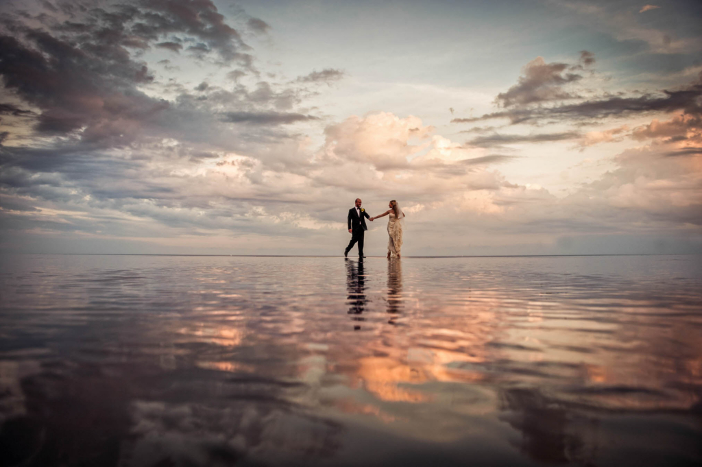 It is always a treat to photograph a wedding in a location that I’ve never shot at before. In 2011 I traveled to the Dominican Republic and photographed a wedding side by side with my husband - something we had never done before and haven’t done since! The bride and groom commissioned both of us to document their day and it was a wonderful wedding from start to finish. After their wedding ceremony on the lawn of their private villa the bride and groom walked with their guests to their reception location at another extraordinarily beautiful home. On the sweeping grounds of that property was an infinity pool that appeared to vanish into the dramatic Dominican Republic sunset. I asked the bride and groom if they’d mind pausing by the pool for a portrait. In order to get the correct perspective to make the photograph look the way I wanted it to I knew that I would have to get down as close to the water’s level as possible. I chose to photograph the scene with my 24-70mm as wide as possible in order to make the bride and groom look small and the space around them look vast. I instructed the couple to simply walk right to left on the size of the infinity pool. I always think that pictures of people pretending to walk look fake and uncomfortable so if I need action in my photographs I want my clients to actually be doing something active! While watching the bride and groom walk from side to side I studied their bodies’ movements so that I would know exactly when to click the shutter. I wanted grace and fluidity in their arms and legs as well as a natural expression on their faces. I also wanted to wait until they were in the middle of the frame so that they would be the central focal point of the image. I don't often put my subjects in the exact center of the frame but I chose to do so in this photograph because I felt that it added an impact to the composition. With the warmth and the drama of the sky above them reflected in the water below them I knew that I had something special! Had I photographed this same portrait from a different height or with a different lens it wouldn’t have resulted in the same final photo. The end result is a fantastic environmental portrait that truly illustrates the beauty of the destination wedding location that the bride and groom chose for their wedding day.