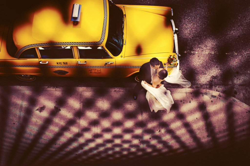 At this 2012 Lighthouse at Chelsea Piers wedding the bride and groom rented a vintage New York taxi to take them around the city for their portraits. They requested some pictures with this cab, and I was only too happy to oblige. Whenever I have a request to take a photograph of the bride and groom in front of an item, vehicle, or building I try to approach it in a slightly new and different way. We had been at the High Line taking photographs of the bride and groom together, and were getting ready to head back over to the Lighthouse for their ceremony. The cab was waiting for us down at the curb. When I looked down on the cab from the High Line staircase, I knew I had an opportunity to make a creative photograph of the scene before me. I placed the bride and groom next to the front of the cab, and I climbed the stairs back up onto the High Line. This image was shot through the metal fence of the High Line. I love it that the pattern of the dots lead your eye directly to the bride, groom, and cab. I was thrilled to be able to give the bride and groom something other than your standard picture of them just standing in front of the car.