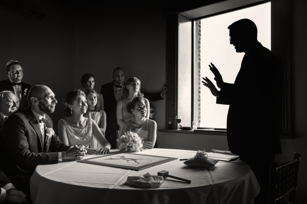 A black-and-white photograph of a wedding ceremony indoors, with a group of people seated around a table listening to an officiant who stands silhouetted by a bright window. The bride and groom sit side by side, smiling and looking toward the speaker. Soft light from the window creates strong contrast, giving the scene a quiet, intimate atmosphere.
