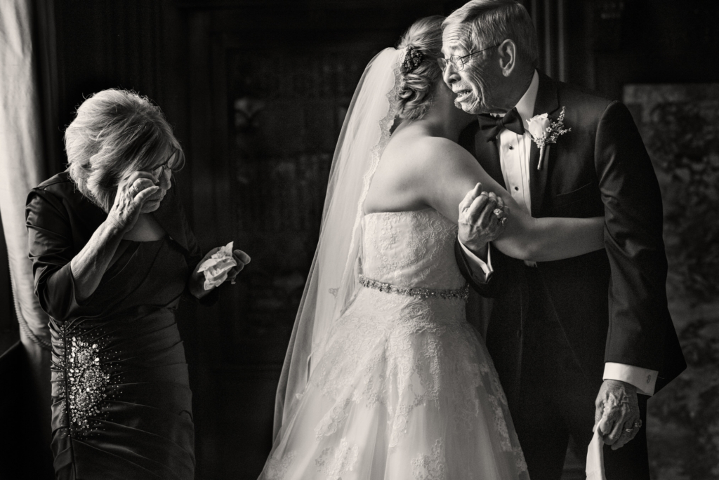 There is not a single thing that I dislike about a Grand Historic Venue wedding. The venue is so perfectly suited for my style of photography. First known as the Tremont Grand, the Grand Historic Venue was built in 1866 as the Grand Lodge of the Maryland Masonic Temple. It was the headquarters for the Maryland Freemasons for over a hundred years, becoming the Tremont Grand in 2005. I am especially crazy about the Library. With it’s dark walls and gorgeous windows, the Library is my ideal setting. I can use the light coming through those windows in dramatic ways, at all hours of the day, in all kinds of weather. The Corinthian Room and the Roman Strada are beautiful spaces as well. On the first floor, the Marble Room is really extraordinary. I also love the Mirror Room and Edinburgh Hall. This venue is extremely convenient, since it attaches directly to the hotel, with great room to start the day off in. This image is especially near and dear to my heart, because it’s a journalistic moment, and we all know how hard those are to capture. You have to find the light, angle yourself perfectly, wait for a moment to occur, and capture it without any interaction with the clients or the scene. When moments like this one happen, they’re a true gift. I mean that photographically, because what wedding photographer wouldn’t want this scene to unfold in front of his or her camera? I also mean that personally, since witnessing such an outpouring of love is really humbling and a huge honor. There are so, so many things that I adore about this image. I love the delicate movement of the mother of the bride as she takes her glasses off to wipe the tears from her eyes. I love that you can’t see the bride’s face, but you can still feel her emotion as she pulls her dad in for a hug. I love every single thing about the father of the bride, from the handkerchief clutched in his hand, to the way he’s holding his daughter’s arm, to the absolutely phenomenal