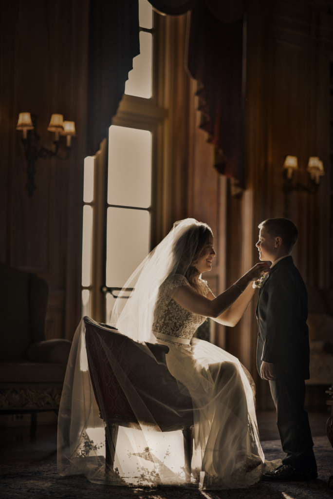 A winter Oheka castle wedding is truly a sight to see. Oheka is majestic enough, with it’s sprawling lawns and pristine architecture. But to see Oheka rising up from the long driveway, snow swirling around it’s exterior and blanketing the great lawns, is truly extraordinary. It takes a brave bride to plan a winter wedding in the northeast, since you never know when a snowstorm will derail your wedding plans! If it all works out, though, and you get a sweet dusting of snow on your wedding day, it really can add something special. This winter wedding at Oheka castle in Huntington, New York, was sweetness from start to finish. The bride and groom are the most lovely, kind, charming people. The venue was intimate and warm, a great contrast to the snowy outdoors. The light was that brilliant winter light, so sharp and brittle, and so very crisp. Right before the ceremony was set to begin, the bride, groom, bridal party, and families were sequestered in the Oheka library to wait. The guests were being seated, and the wedding ceremony was about to start. The bride was sitting in the most brilliant patch of light, that crystalline-perfect winter light that I adore so much. She was adjusting the bow tie of the little ring bearer, and the scene was perfect.