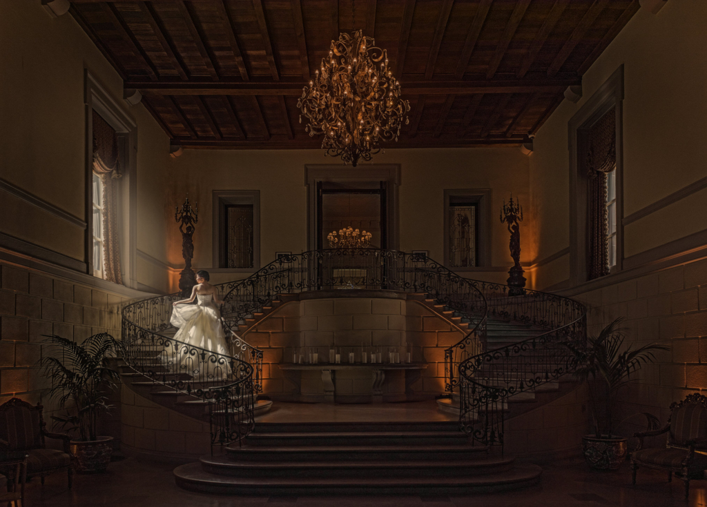 A bride stands alone on a grand, curved staircase inside an elegant historic hall. Warm chandelier light fills the room, highlighting ornate railings, tall walls, and rich wooden ceilings. The scene feels dramatic, cinematic, and quietly contemplative.