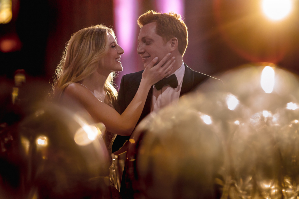 A couple dressed in formal attire smile at each other under soft, golden stage lights. The woman gently cups the man’s face as blurred glassware and glowing lights frame them in the foreground, creating a romantic, cinematic atmosphere.
