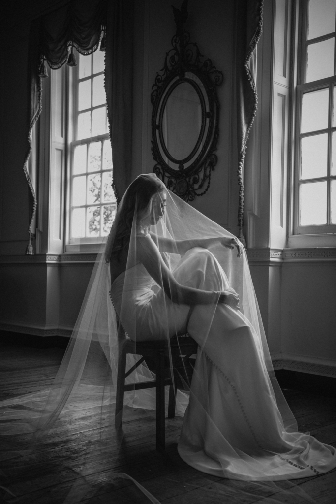 A bride sits quietly on a chair in an elegant room, her long veil draped over her shoulders and flowing around her. Soft window light outlines her figure, creating a calm, reflective mood against ornate mirrors and tall windows.