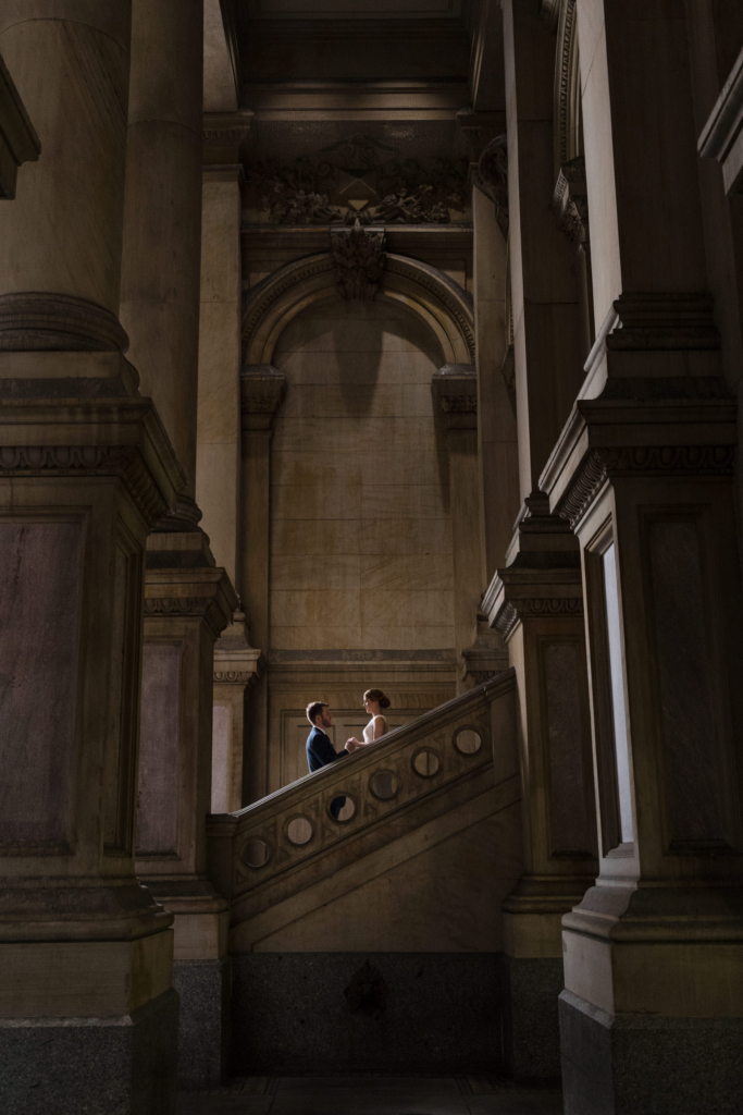 A bride and groom stand facing each other on a wide stone staircase, framed by towering columns and classical architectural details. Soft, moody light emphasizes the scale of the space, making the couple appear small and intimate within the monumental interior.