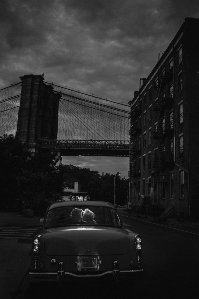 A black-and-white image of a couple sitting closely together in the front seat of a vintage car at night. In the background, a large suspension bridge rises between dark buildings, creating a cinematic urban scene.