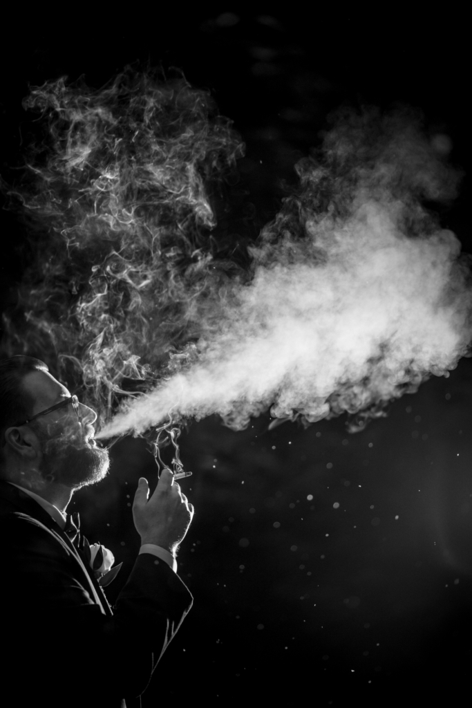 A black-and-white close-up of a man in formalwear exhaling a cloud of smoke into the dark air. The smoke curls and drifts dramatically in the light, forming soft, abstract shapes around his face.