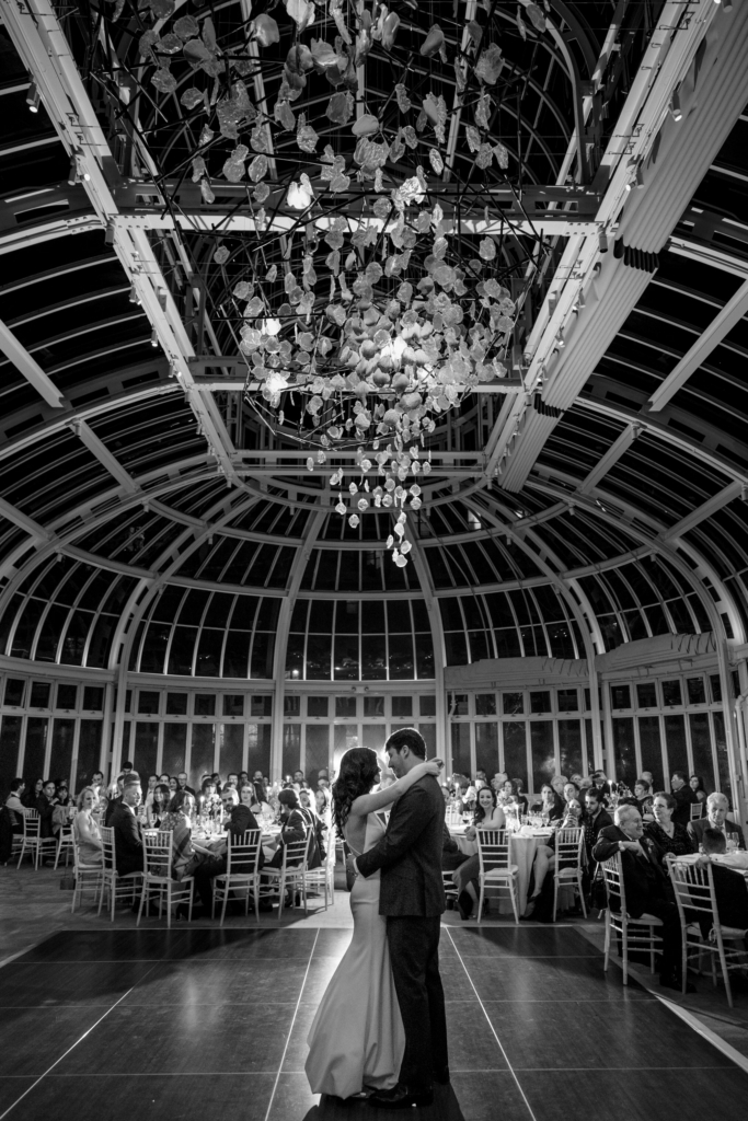 A bride and groom share their first dance at the center of a grand glass-roofed hall, surrounded by seated guests. Above them, a large chandelier-like installation hangs from the ceiling, creating a dramatic, elegant atmosphere.