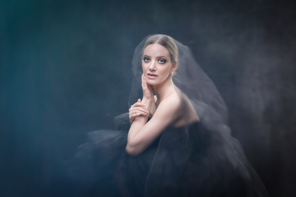 A woman faces the camera with a soft, calm expression, her hand resting gently against her cheek. She is wrapped in a translucent veil or fabric that appears to flow around her, creating a dreamy, ethereal atmosphere against a dark background.