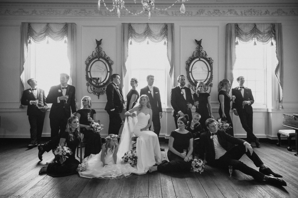 A bride sits centered in a formal room with tall windows, ornate mirrors, and a chandelier, surrounded by bridesmaids and groomsmen in coordinated formal attire. Some stand, others sit or lounge on the floor, creating a relaxed yet stylish group portrait with soft, natural light.