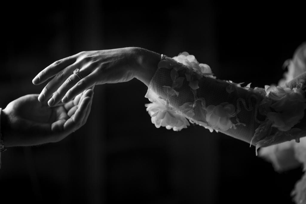 In a dramatic black-and-white close-up, two hands reach toward each other in mid-air. One hand wears a ring and emerges from a sheer, floral-textured sleeve, the light catching delicate details while the background fades into darkness.