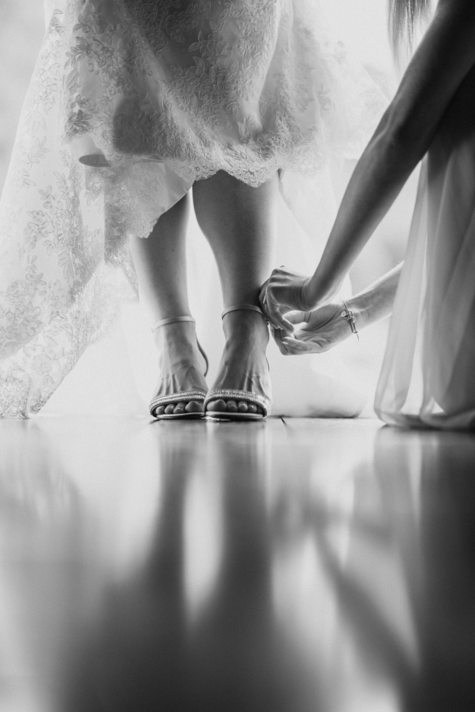A close-up view of a bride’s feet in elegant heels beneath the lace hem of her dress, reflected softly on the floor. Another person kneels to fasten the ankle strap, capturing a quiet, intimate moment of preparation before the ceremony.