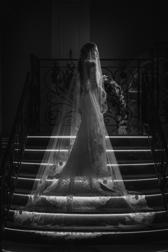 A black-and-white photograph of a bride standing on illuminated stairs, her long veil flowing behind her. She holds a bouquet and is softly lit from the side, creating a dramatic, elegant silhouette in a dark interior space.