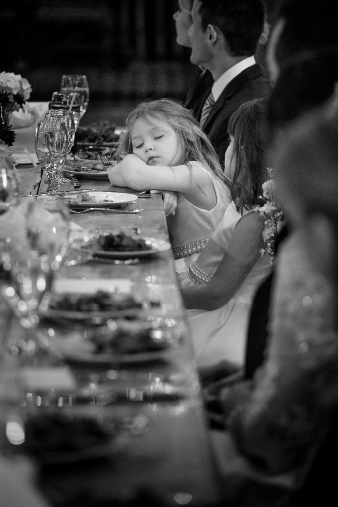 A young girl in a dress has fallen asleep at a long dinner table, resting her head on her folded arms. Plates, glasses, and adults seated around her suggest a formal celebration continuing late into the evening.