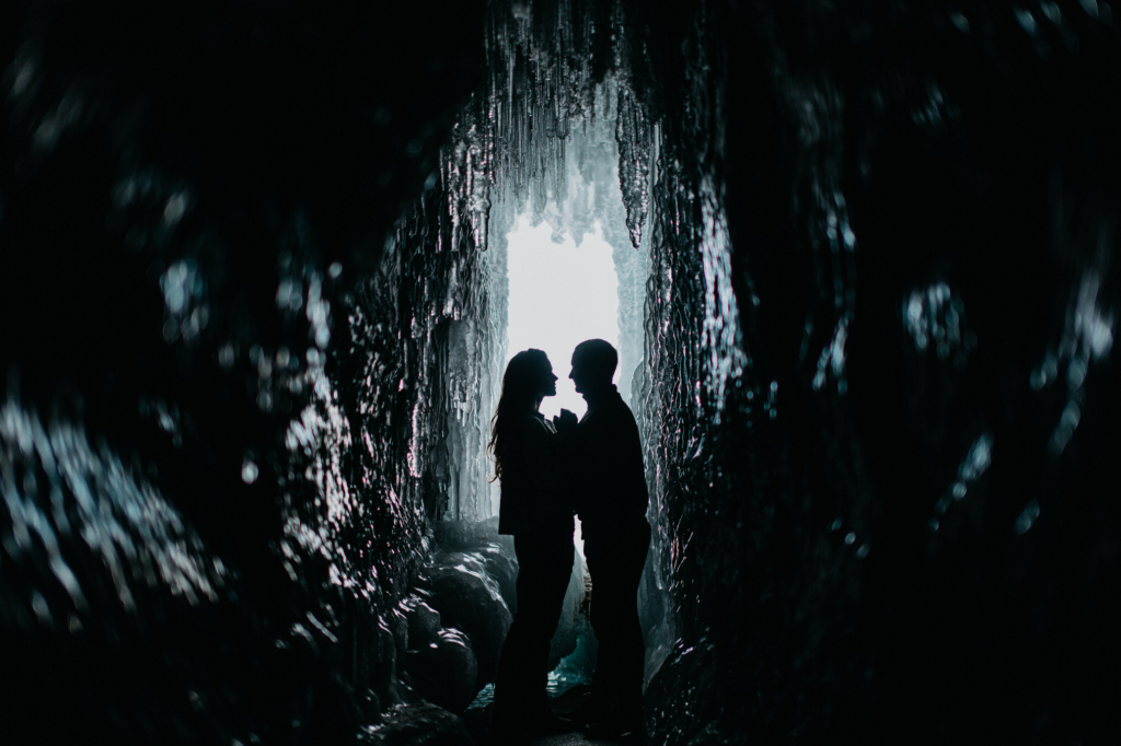 A silhouetted couple stands facing each other inside an icy cave. Jagged ice walls surround them, and bright white light pours in from the cave opening behind, framing their figures in darkness and creating a dramatic, intimate scene.
