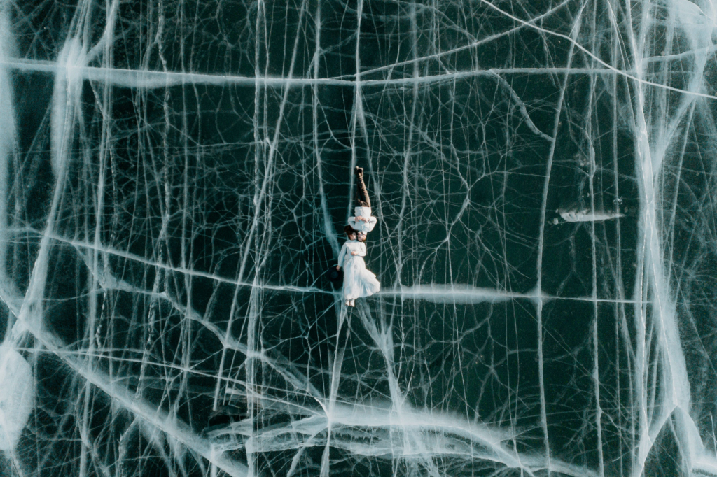 A couple lies side by side on a frozen lake, seen from directly above. The dark ice beneath them is crossed with white cracks that form a web-like pattern, while their light clothing contrasts sharply against the textured surface, creating a quiet, surreal sense of stillness.