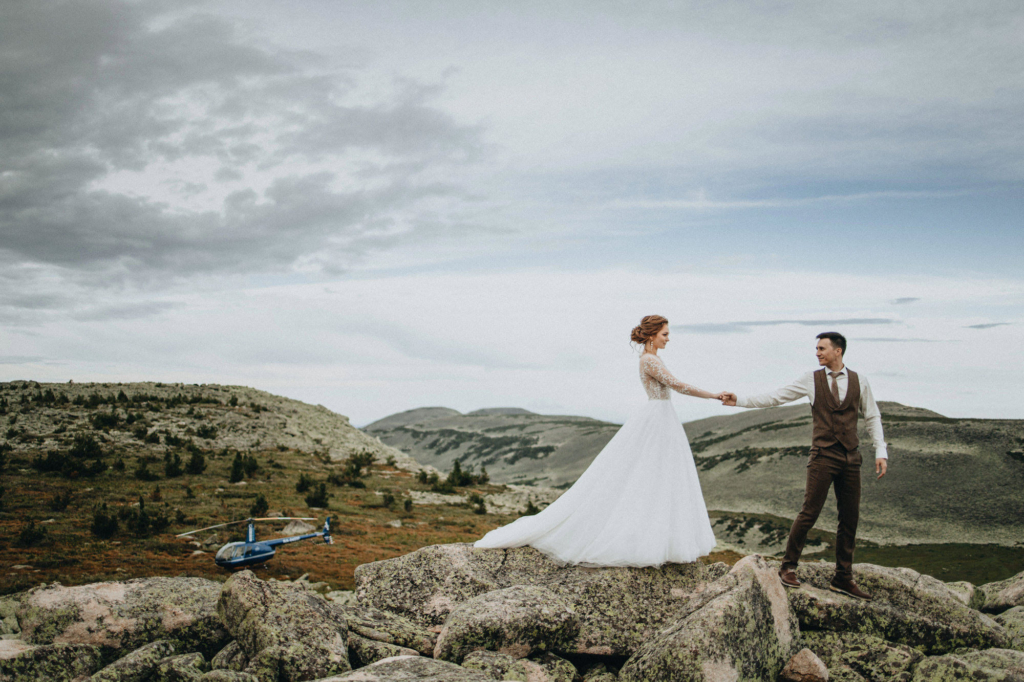 A bride and groom stand on separate rocks in a wide mountain landscape, holding hands across the gap between them. The bride wears a long white dress, the groom a vest and trousers. In the background, rolling hills stretch under a cloudy sky, with a small helicopter resting on the ground nearby.