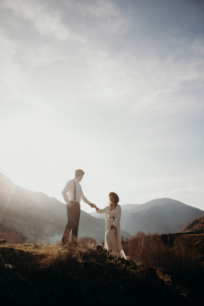 A couple stands on a rocky hillside, holding hands against a bright sky. The man wears suspenders and a white shirt, the woman a long light dress and a hat. Mountains fade into the background, and soft sunlight creates a calm, intimate atmosphere.