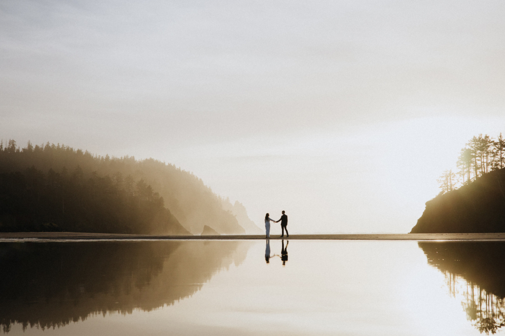 A couple stands holding hands on a wide, quiet beach. Their reflections are mirrored in shallow water beneath them. Misty forested cliffs rise on both sides, and soft light fills the open sky, creating a calm, spacious atmosphere.