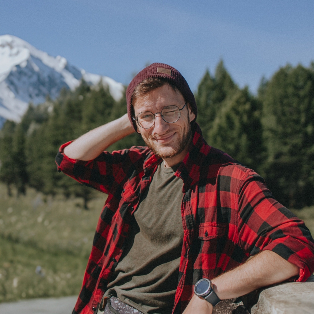 “An image of Georgy Shishkin wearing glasses and a red hat, with snow-covered mountains in the background.”