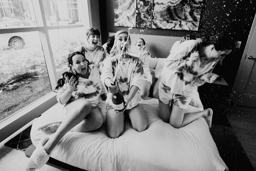 Eternal Love: Capturing the High School Sweetheart's Wedding Day at Visit Arts Rockville
A group of women sits and kneels on a bed in a hotel room, laughing and reacting as a bottle of sparkling wine sprays into the air. The image is in black and white, capturing motion as droplets scatter across the scene. Their expressions and body language convey surprise, joy, and playful celebration.